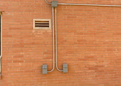 Conduit pipes running along a brick wall with electrical boxes and a small vent.